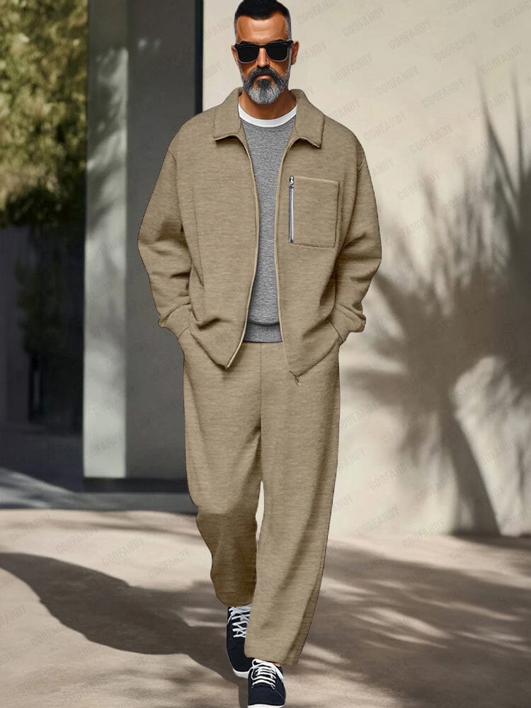 Man wearing a beige outfit with sunglasses standing against a light-colored wall.