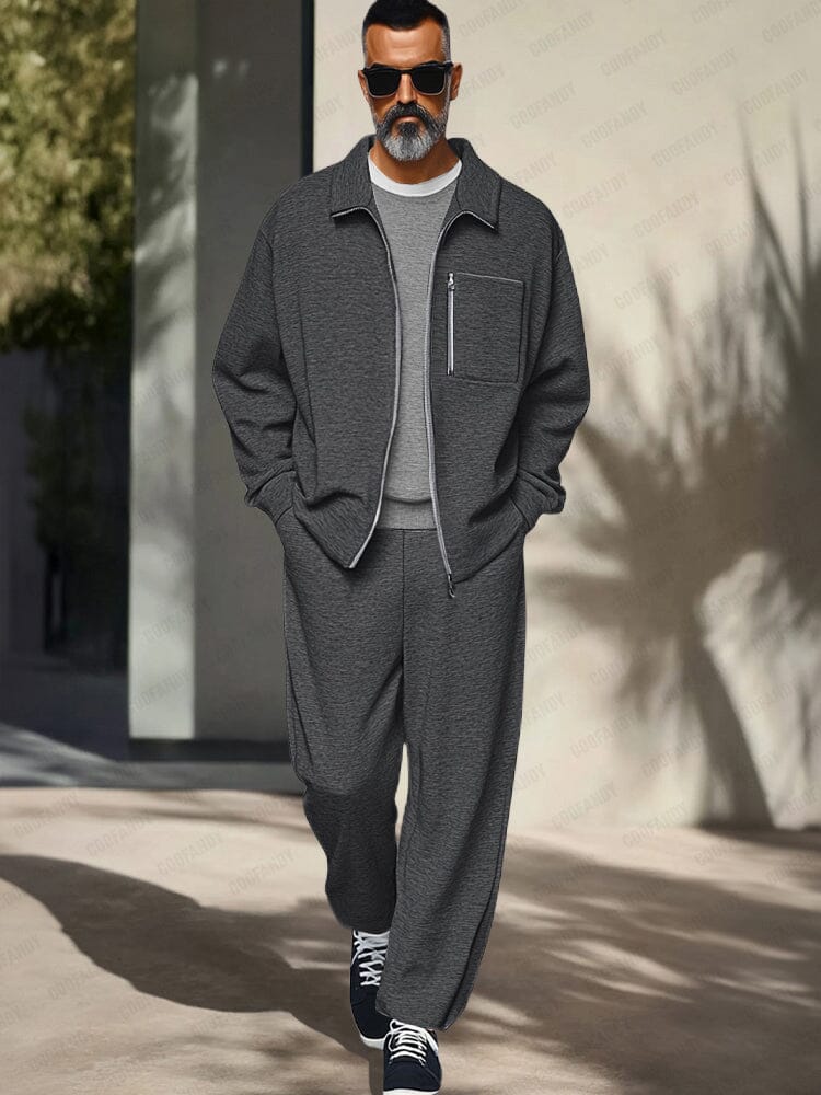 Man wearing a gray outfit with sunglasses standing against a light-colored wall.