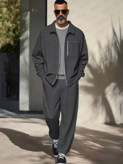 Man wearing a gray outfit with sunglasses standing against a light-colored wall.