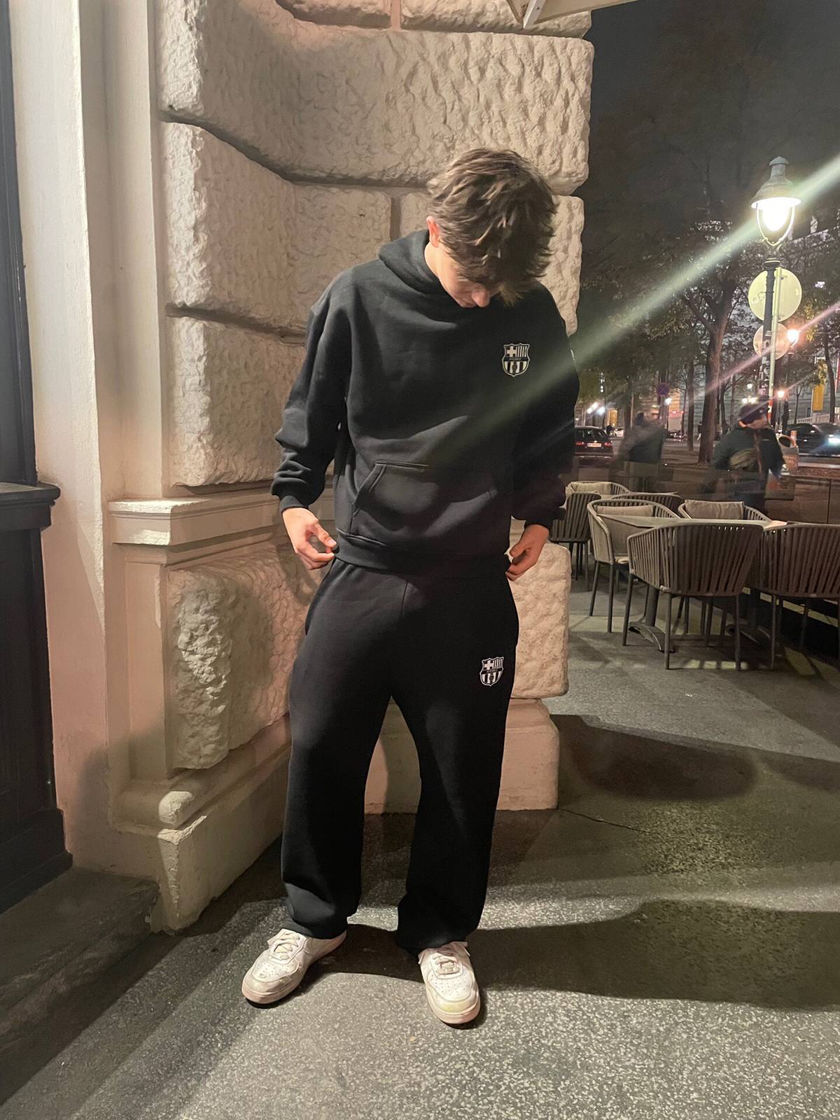 Person wearing a black hoodie and pants with a logo, standing on a street at night.