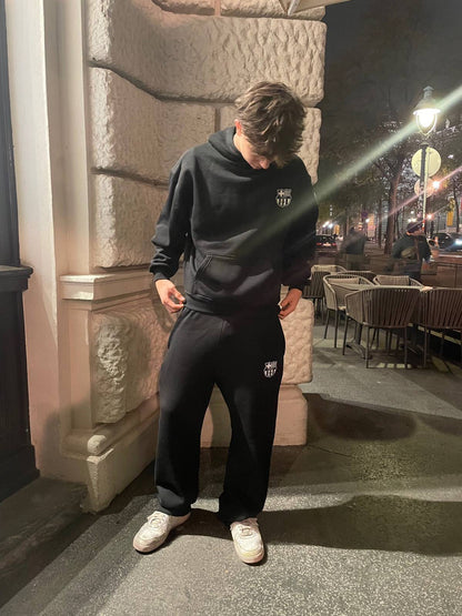 Person wearing a black hoodie and pants with a logo, standing on a street at night.