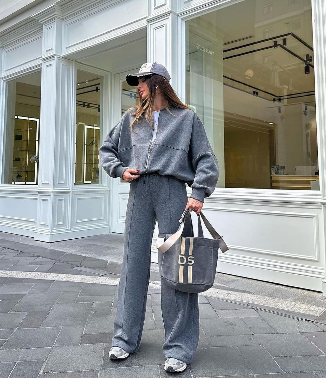 Person wearing a gray tracksuit with a cap, standing in front of a store.
