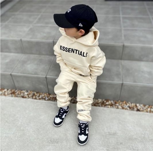 Child wearing a beige 'ESSENTIALS' hoodie and pants with a black cap, standing on a tiled floor.