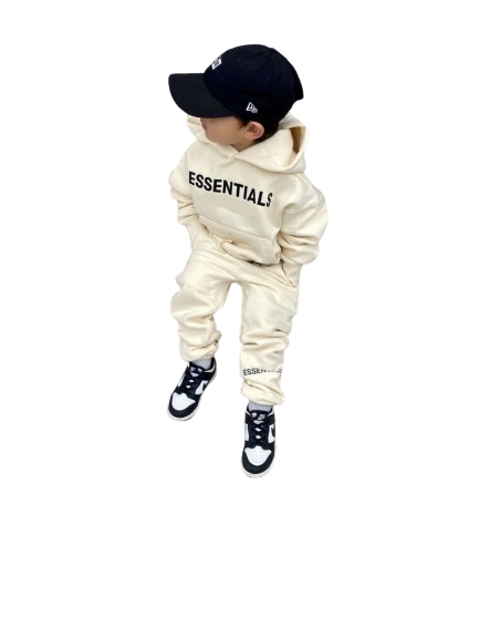 Child wearing a cream hoodie and pants set with 'ESSENTIALS' text, black cap, and shoes on a white background