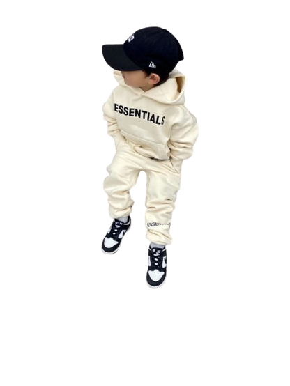 Child wearing a cream hoodie and pants set with 'ESSENTIALS' text, black cap, and shoes on a white background