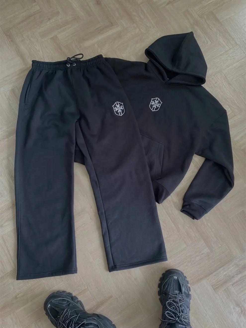 Black tracksuit with logos on pants and jacket laid out on a wooden floor.