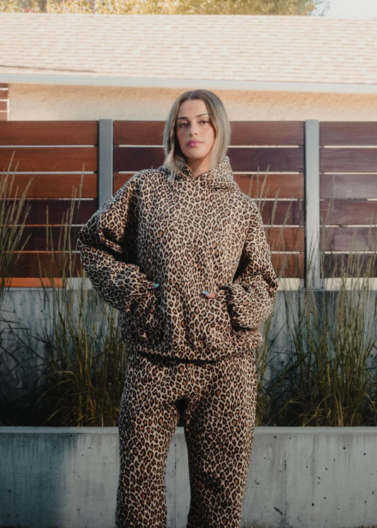 Person wearing a leopard print hoodie and pants standing outdoors with a wooden fence and grass in the background.