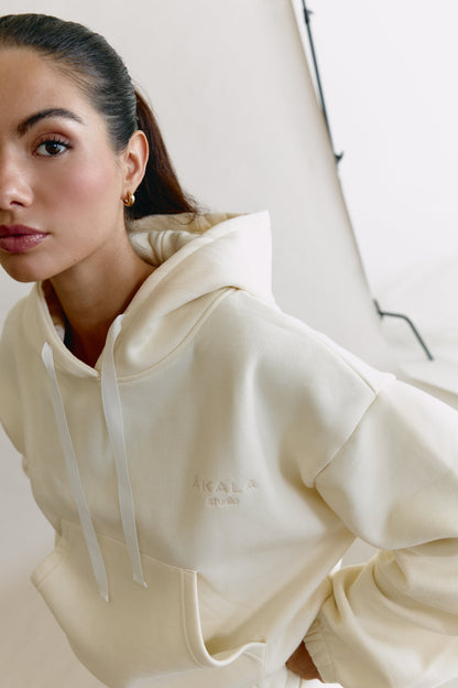 Woman wearing a cream-colored hoodie with a brand logo on a neutral background