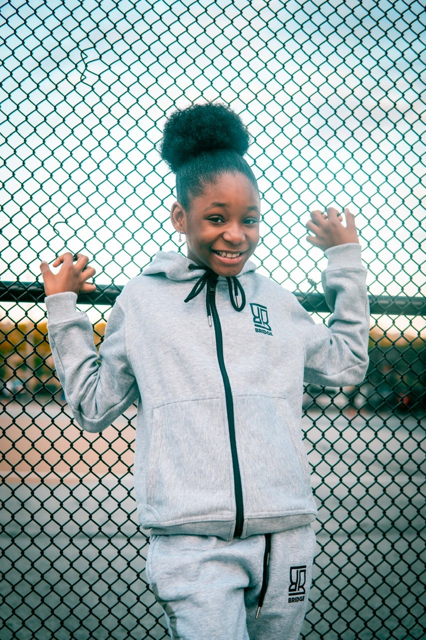 Child wearing a gray tracksuit with a logo in front of a chain-link fence.