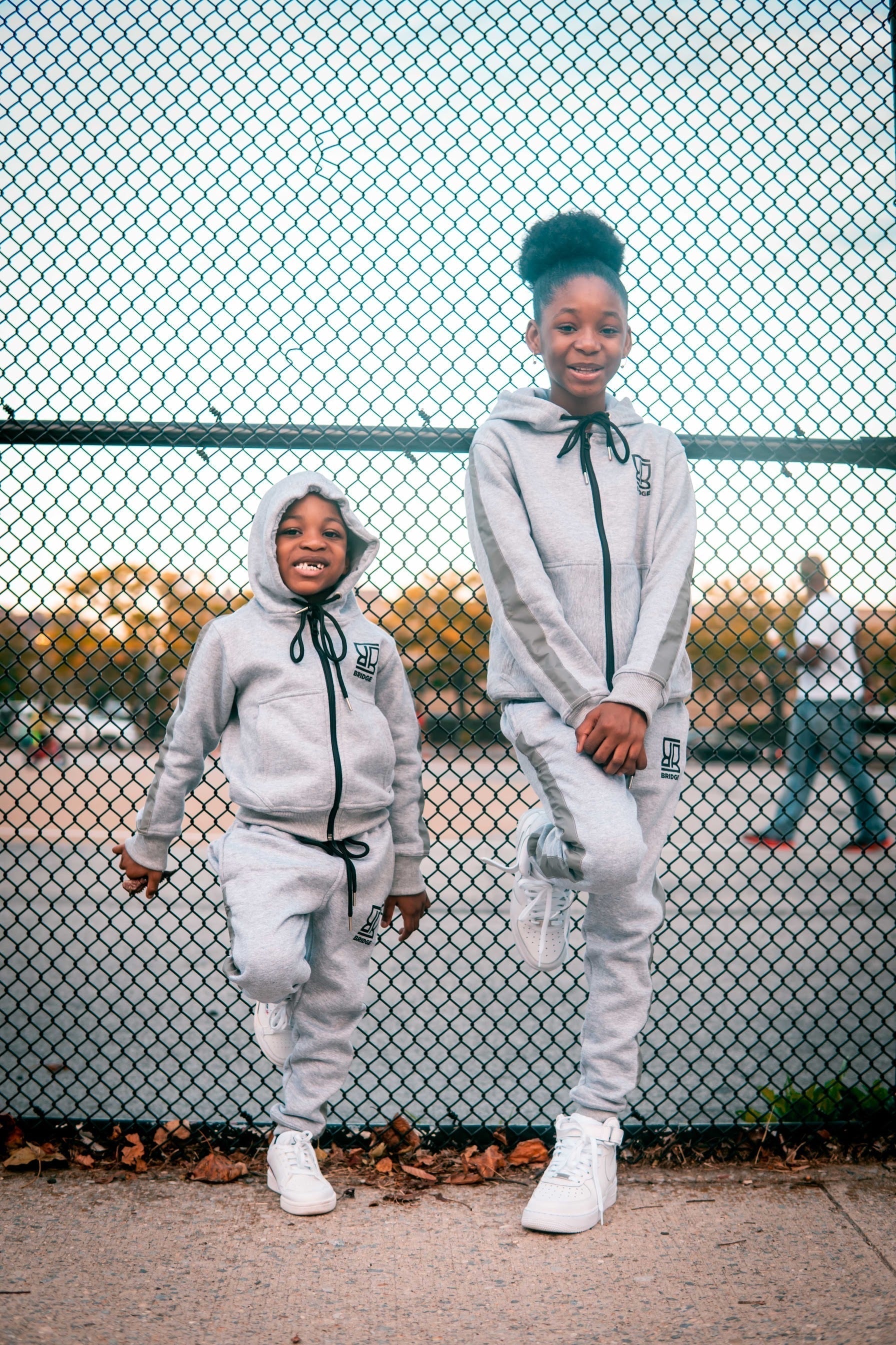 Two children in matching gray outfits standing in front of a chain-link fence.