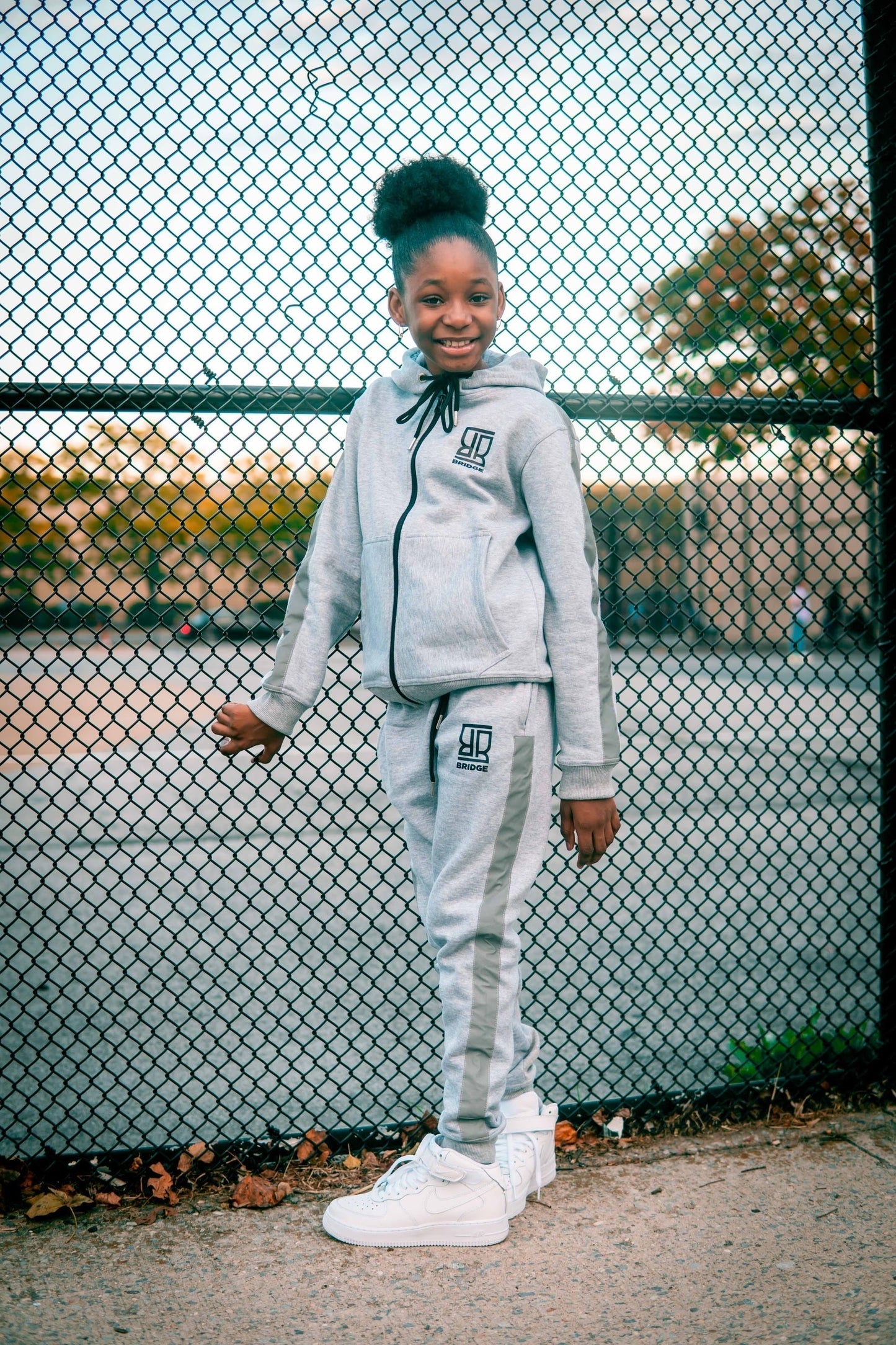 Child wearing a gray tracksuit with a logo, standing in front of a chain-link fence.