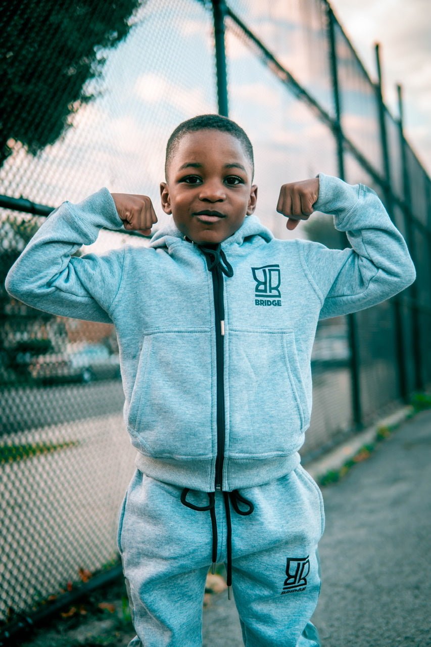 Child wearing a light blue tracksuit with a logo, flexing muscles outdoors.