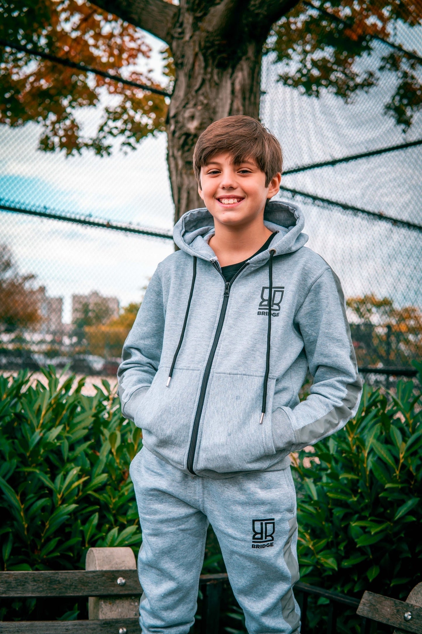 Young boy wearing a gray hoodie and pants with a logo, standing outdoors.