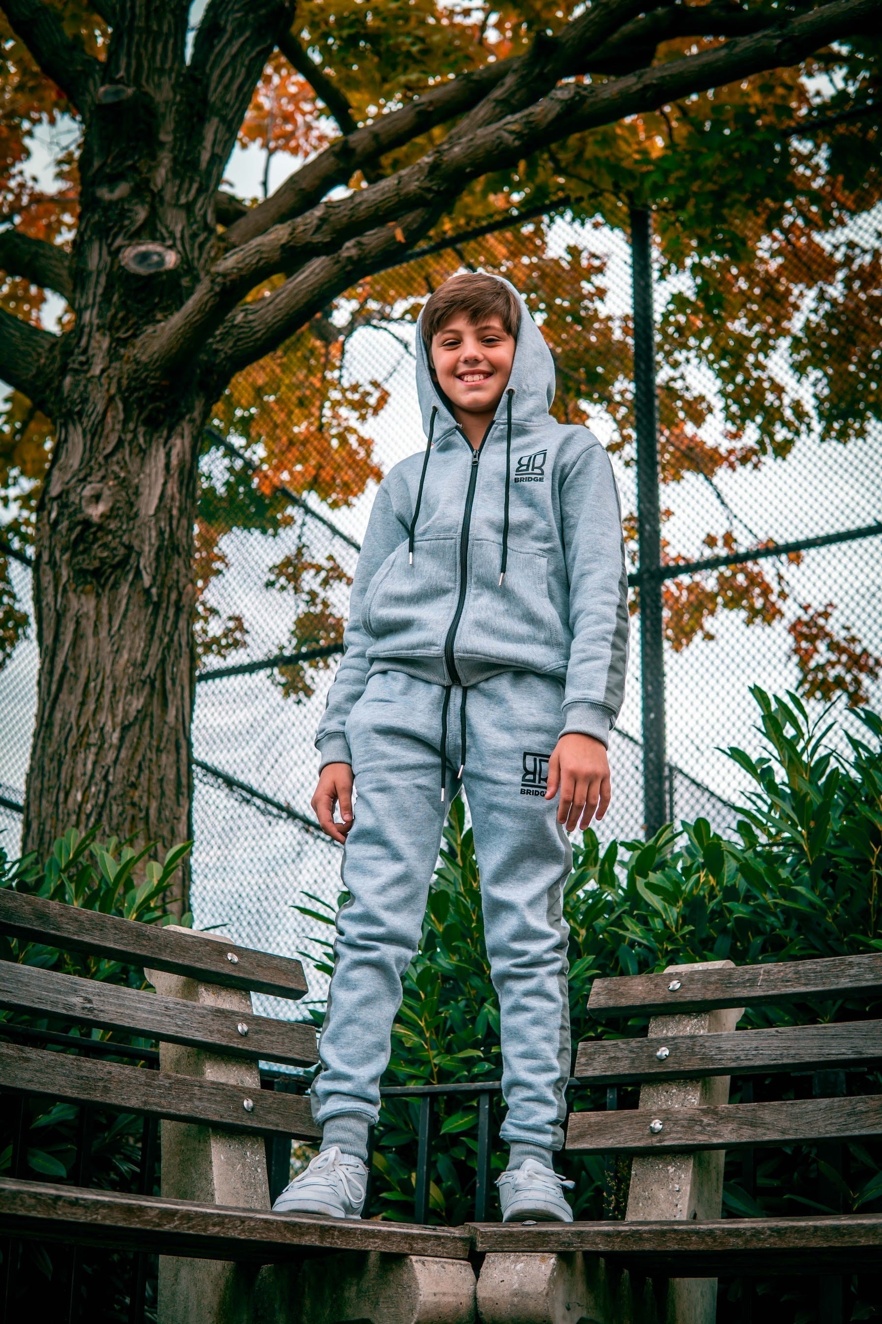 Child wearing a gray hoodie and pants standing on a wooden bench with trees and foliage in the background