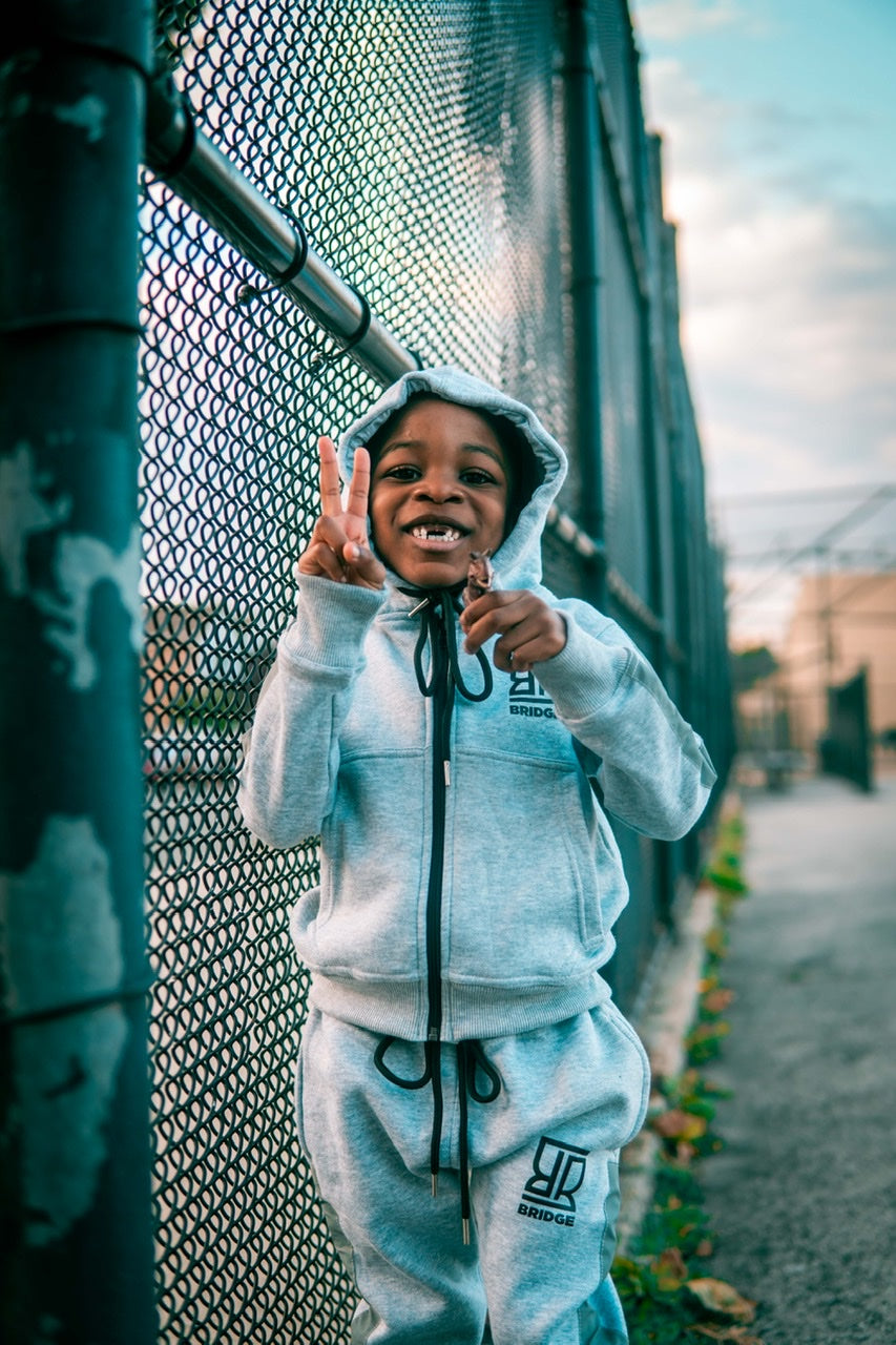 Child in a white hoodie and pants standing next to a chain-link fence.