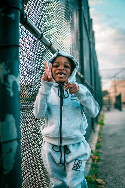 Child in a white hoodie and pants standing next to a chain-link fence.