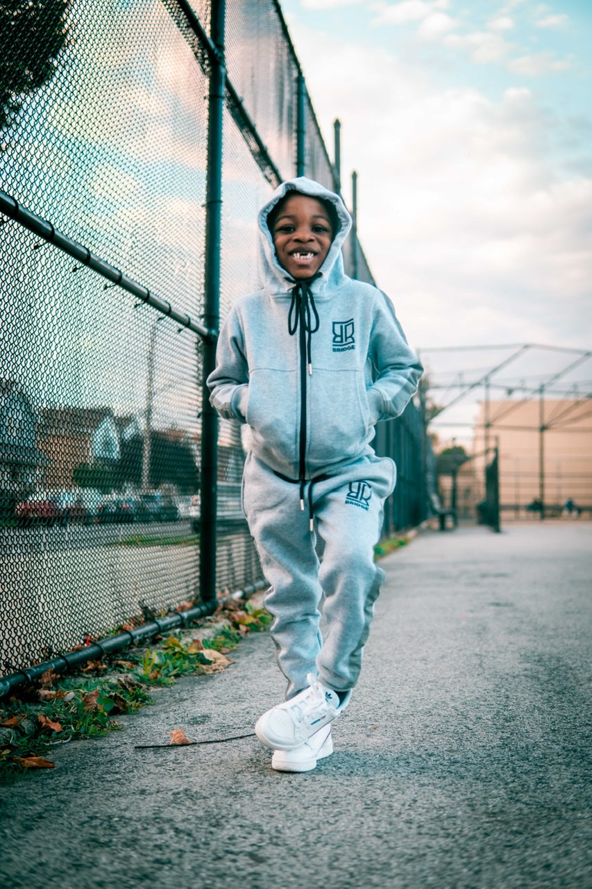 Child wearing a gray hoodie and pants with a logo, standing on a paved area near a chain-link fence.