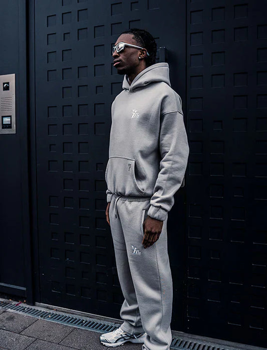 Person wearing a gray hoodie and pants standing against a dark textured wall.